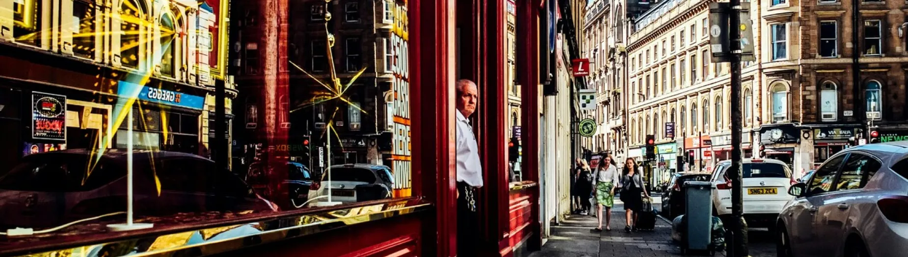 A bustling city street with historic buildings, a man stands in a doorway. Cars are parked along the road, and pedestrians walk on the sidewalk. Reflections are visible in the storefront window.